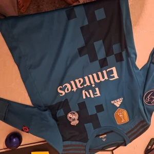 Real Madrid Ronaldo långärmad tröja - Säljer en officiell Real Madrid långärmad fotbollströja från Adidas, med Ronaldo 7 på ryggen. Tröjan är blå med mörkblå pixelmönster, broderat klubbmärke, Champions League-märke och FIFA World Champions 2017 badge. Materialet är lätt och andas, perfekt för fotbollsfans.