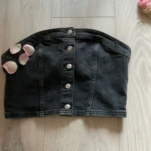 Svart jeans tubtopp med knappar - Cool svart tubtopp i jeansmaterial med silvriga knappar framtill. Toppen har markerade sömmar som ger en snygg form och är perfekt för dig som gillar en edgy stil. Passar dig som vill sticka ut och köra på en trendig look.