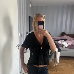 Svart stickad kofta med kort ärm - Kortärmad svart stickad kofta med två stora knappar framtill och fickor. Djup v-ringning och croppad passform som ger en chill vibe. Perfekt att slänga över ett linne eller t-shirt för en snygg lager-på-lager-look.