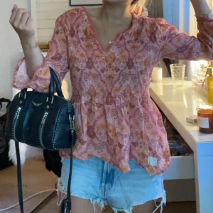 Rosa blommig blus med v-ringning - Supersöt blommig blus från Odd Molly, i rosa och orange toner med v-ringning och långa ärmar.Blusen har en flowy passform och volangdetaljer vid ärmslut och nederkant. Perfekt för dig som gillar boho-vibe och vill ha något färgglatt och bekvämt.