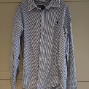 Randig skjorta från Ralph Lauren - Klassisk blåvit randig skjorta från Ralph Lauren med lång ärm och knappar framtill. Skjortan har en snygg krage och den ikoniska broderade loggan på bröstet. Perfekt för dig som gillar stilrena och tidlösa plagg.