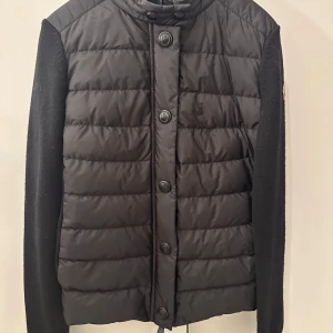 Svart pufferjacka från Moncler - Säljer en svart pufferjacka från Moncler  i storlek S. Jackan har stickade ärmar, quiltad front med knappar och hög krage. Klassisk Moncler-logga på ärmen. Perfekt för dig som vill ha en stilren och varm jacka med exklusiv känsla.