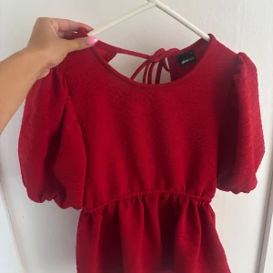 Röd blus från Gina Tricot - Supersöt röd blus med puffärmar och knyt i ryggen från Gina Tricot. Blusen har en lätt struktur i tyget, markerad midja och öppen rygg med två knytband. Perfekt för dig som vill sticka ut med en färgstark look! 
