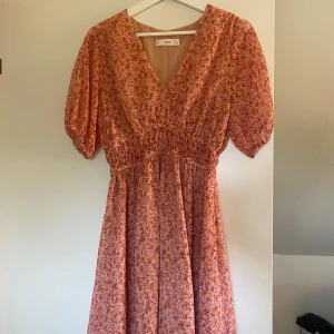 Blommig rosa klänning från Mango - Supersöt rosa klänning från Mango med småblommigt mönster i rött och gult. Klänningen har v-ringning, korta puffärmar och smock i midjan för en snygg passform. Perfekt för sommarens alla tillfällen!