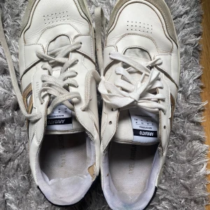 Vita sneakers från Axel Arigato - Snygga vita sneakers från Axel Arigato med klassisk siluett och rund tå. Skorna har snörning, detaljer i beige mocka och robust sula med tydlig logga undertill. Tillverkade i skinn och textil, perfekta för en clean och stilren look.  Hälkappan på vänstra skon har slitits o förstörts i tvätt, skriv gärna för mer info…
