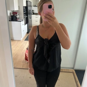 Svart linnes med stor rosett - Säljer en svart linnetopp med tunna axelband och en stor rosett framtill. Toppen har en lätt glansig finish upptill och är i ett tunt, flowy material som känns lyxigt. Perfekt för dig som gillar en chic och trendig look med det lilla extra. Aldrig använd