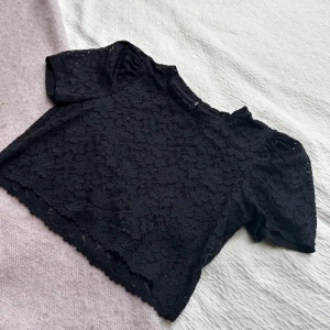 Svart spetstopp - Säljer en svart croppad blus med korta puffärmar och blommigt spetsmönster. 