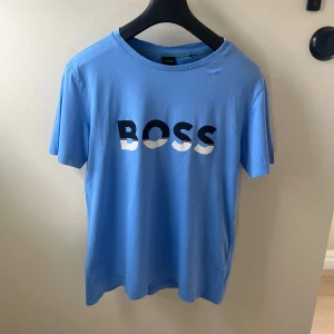 Blå BOSS t-shirt i bomull - Snygg blå t-shirt från BOSS med stor logga framtill i svart och vitt. Klassisk rund halsringning och korta ärmar. Tillverkad i mjuk bomull som känns skön mot huden. Perfekt för dig som gillar stilrena och trendiga plagg.