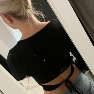 Svart croppad topp med knytband - Säljer en svart croppad topp med långa ärmar och snygg snörning i ryggen. Toppen har en tight passform och är perfekt att styla med höga jeans. Enkel men trendig design som passar till många olika looks.