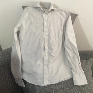 Randig skjorta från Morris, strl 41 - Snygg vit skjorta med tunna svarta ränder från Morris. Klassisk krage, långa ärmar och knäppning framtill. Tillverkad i bomullstyg från Thomas Mason, vilket ger en lyxig känsla. Perfekt för dig som gillar stilrena och tidlösa plagg.