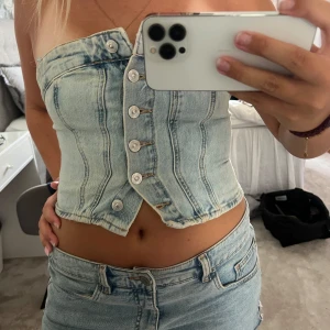 Ljusblå jeans korsett med knappar - Säljer en snygg ljusblå jeanskorsett med markerade sömmar och cool knappstängning framtill. Toppen är axelbandslös och croppad, perfekt till sommarens outfits. Ger en trendig och edgy vibe!