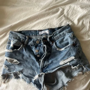 Blå slitna jeansshorts från Zara - Säljer ett par blå jeansshorts från Zara med råa kanter och slitna detaljer. Shortsen har knappar framtill och klassisk femficksdesign. Perfekta för sommaren om du gillar en avslappnad och edgy stil.