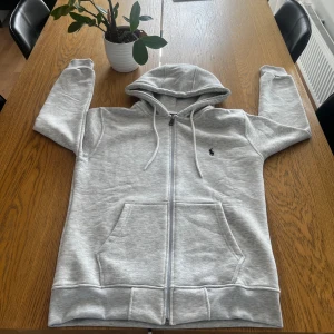 Grå zip hoodie ifrån Polo Ralph Lauren - Snygg ljusgrå hoodie från Polo Ralph Lauren med dragkedja, huva med snörning och klassisk broderad logga på bröstet. Tröjan har känguruficka framtill och är tillverkad i mjuk bomullsmix. Perfekt för chill dagar eller när du vill ha en clean streetstyle.
