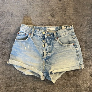 Jeansshorts från Zara - Snygga ljusblå jeansshorts från Zara med slitna detaljer. Klassisk femficksmodell med knappar framtill och bälteshällor. Perfekta för sommaren och ger en avslappnad vibe.