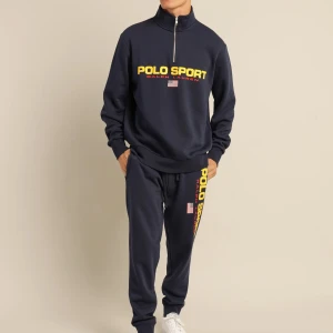 Mörkblå dress från Ralph Lauren  - Snygg mörkblå långärmad tröja och byxor från Polo Sport Ralph Lauren med gul logga och amerikansk flagga på bröstet. Tröjan har half zip, hög krage och ribbade muddar. Perfekt för en sportig och avslappnad stil.