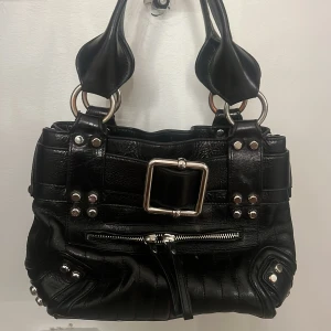svart vintage skinnväska från Karen Millen - svart vintage handväska i skinn från Karen Millen med stora silvriga metalldetaljer, markerat spänne och nitar. väskan har dubbla handtag, dragkedja framtill och flera fack inuti. väskan är köpt för 4000kr. pris kan diskuteras!💞