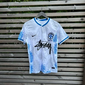 Vit Brasilien fotbollströja Stüssy x Nike - Snygg vit Brasilien fotbollströja från Nike i samarbete med Stüssy. Tröjan har blå detaljer, palmbladsmönster i blått och grått, broderat CBF-märke och Stüssy-logga framtill. Tillverkad i Dri-FIT-material som andas. Perfekt för dig som vill sticka ut på planen.