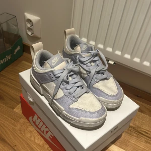 Nike Dunk Low i vitt och lila - Snygga Nike Dunk Low sneakers i vitt och lila med klassisk siluett. Skorna har snörning, platt sula och är tillverkade i en mix av skinn och syntet. Perfekta för dig som gillar streetstyle och vill ha en clean look. Använda men enkelt att tvätta rent då de tyg och ”läder”