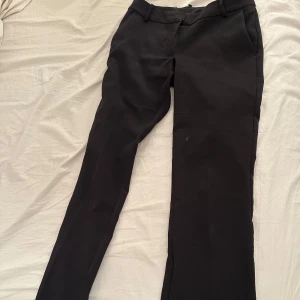 Svarta kostymbyxor med raka ben - Snygga svarta kostymbyxor med klassisk utsvängd passform och diskreta fickor i sidorna. Perfekta för en clean och stilren look. Byxorna har hällor för bälte och stängs med dragkedja och knapp.