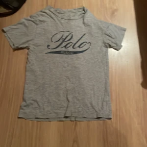 Grå Polo Ralph Lauren t-shirt - Grå t-shirt från Polo Ralph Lauren med stor blå logga framtill. Klassisk passform med korta ärmar och rund hals. Tillverkad i mjuk bomull, perfekt för en chill och stilren look.