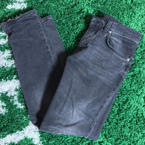  Nudie Jeans - Säljer ett par grå jeans från Nudie Jeans med klassisk femficksdesign och svarta broderade detaljer på bakfickan. Jeansen har en rak passform och är tillverkade i mjukt bomullsjeans-tyg. Perfekta för dig som gillar en stilren och avslappnad look, Grå/svart slim fit