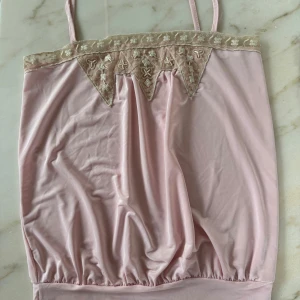 Rosa linnetopp med spetsdetaljer - Söt ljusrosa linnetopp med smala axelband och beige spets med broderade blommor upptill. Toppen har en lätt glansig yta och ett avslappnat fall med resår nertill. Perfekt för dig som gillar romantiska detaljer och mjuka pasteller.