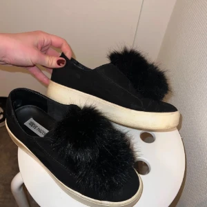 Svarta sneakers med päls från Steve Madden - Svarta slip-on sneakers från Steve Madden med fluffig svart pälsdetalj på ovansidan. Skorna har en vit platt sula och är tillverkade i mocka med pälsdetalj som ger en unik och trendig look. Skorna har lite missgärningar på sulan men inget man tänker på när man har på sig skorna!