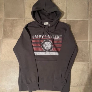 Saint Laurent Hoodie - Tja, säljer en eftertraktad Saint Laurent Hoodie som är i super bra skick. Tröjan är i brun/grå och är i storlek M och har du frågor är de bara att höra av sig✌️