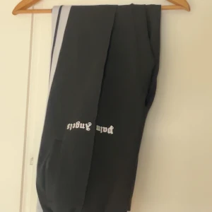 Palm Angels svarta mjukisbyxor - Svarta mjukisbyxor från Palm Angels med vita sidoränder och logga i vitt tryck på benet. Byxorna har en sportig vibe och är tillverkade i ett mjukt syntetmaterial. Perfekta för en chill och trendig streetwear-look.