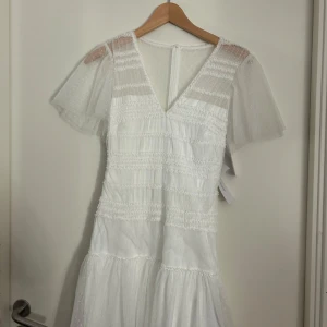 Vit långklänning med volang och mesh - Superfin vit långklänning med v-ringning, volangdetaljer och transparenta meshärmar. Klänningen har diskret prickigt mönster i tyget och en romantisk känsla. Perfekt för dig som vill ha en luftig och söt look med extra detaljer.