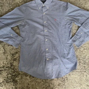 Blårutig skjorta från Polo Ralph Lauren - Snygg blårutig skjorta från Polo Ralph Lauren med klassisk button-down krage och lång ärm. Skjortan har små vita och blå rutor, vita knappar och den ikoniska rosa broderade loggan på bröstet. Perfekt för dig som gillar stilrena och tidlösa plagg. Storlek S, hör av dig vid fundering