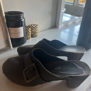 Bruna trätofflor från Moheda - Klassiska bruna trätofflor från Moheda med bred rem i skinn och stor metallspänne. Snyggt handgjorda med robust träsula och låg klack. Perfekta för dig som gillar en retro och genuin stil med svensk design.