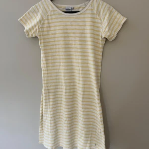 Randig kortklänning från Ebbe - Supersöt kortklänning från Ebbe i vitt och ljusgult med smala horisontella ränder. Klänningen har rund halsringning och korta ärmar, perfekt för varma dagar. Tillverkad i mjuk bomull som känns skön mot huden.