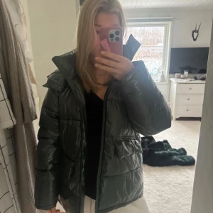 Grön pufferjacka från Minus - Säljer en glansig grön pufferjacka från Minus i storlek 42. Jackan har hög krage, dragkedja framtill och två stora fickor. Perfekt för kalla dagar och har en oversized vibe. Materialet är syntet och jackan är quiltad för extra värme. Jag har oftast storlek 40 men passar bra för mig också. Nypris 2000