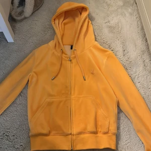 Gul velour hoodie från Juicy Couture - Superfin gul hoodie från Juicy Couture i mjuk velour med dragkedja framtill, snörning i huvan och broderad JC-logga på bröstet. Perfekt för dig som vill ha en chill och trendig look. Känguruficka framtill och ribbade muddar. Har aldrig använt koftan. 