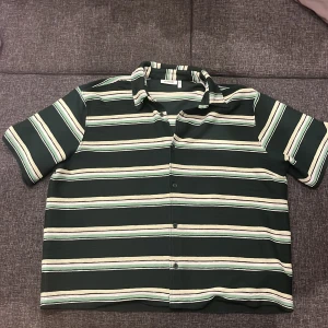 Randig skjorta från Weekday, grön/vit - Nypris 400. Snygg randig skjorta från Weekday i grönt, vitt och ljusgult. Skjortan har korta ärmar, klassisk krage och knappar framtill. Perfekt för dig som gillar en chill och stilren look. Materialet är mjukt och känns skönt mot huden.