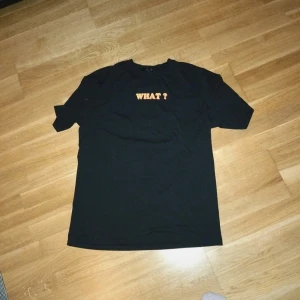 Svart t-shirt med orange text - Svart t-shirt med trycket 'WHAT?' i orange på bröstet. Klassisk passform med rund hals och korta ärmar. Perfekt för dig som gillar enkla men uttrycksfulla plagg.