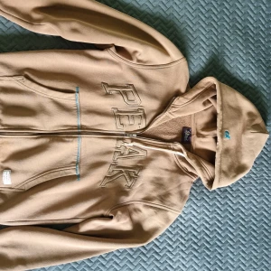 Beige hoodie från Peak Performance - Snygg beige hoodie från Peak Performance med broderad logga över bröstet och dragkedja framtill. Tröjan har huva, långa ärmar och praktiska fickor. Perfekt för en avslappnad och sportig stil.