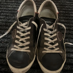 Svarta sneakers från Philippe Model - Svarta sneakers från Philippe Model med vita snören och vit sula. Skorna har detaljer i mocka och skinn, samt en vit hälkappa med röd söm. Sidan pryds av en sköldformad patch med röd kant. Perfekta för en avslappnad streetstil.