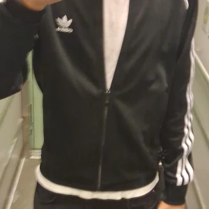 Svart Adidas track jacket med vita ränder - Svart Adidas track jacket med klassiska vita ränder längs ärmarna och broderad logga på bröstet. Jackan har dragkedja framtill, ribbade muddar och är tillverkad i glansig polyester. Perfekt för en sportig och avslappnad stil. Kan diskutera om priset det är bara att kontakta