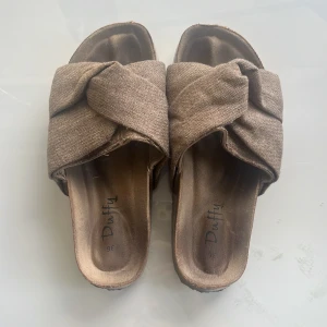 Beige sandaler med knut från Duffy - Snygga beige sandaler från Duffy med breda korslagda band i textil och cool knutdetalj upptill. Skorna har platt sula och öppen tå, perfekta för varma dagar. Stilren och enkel design som passar till många outfits.
