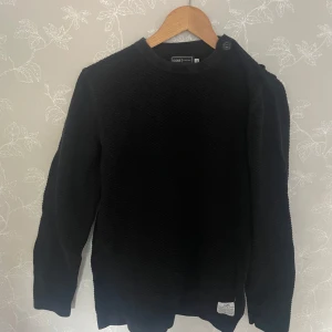 Svart stickad tröja Jack & Jones - Svart stickad tröja från Jack & Jones Core med diskret strukturmönster. Rund halsringning, knappdetalj vid axeln och ribbade muddar. Perfekt för dig som gillar en enkel men snygg look.