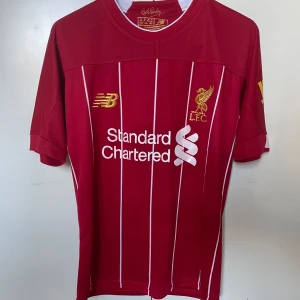 Liverpool Salah 11 matchtröja New Balance - Liverpool FCs officiella matchtröja i röd med vita tunna ränder, tryckta sponsorloggor och Salah 11 på ryggen. Tillverkad i lätt och ventilerande polyester, klassisk passform och korta ärmar. Guldiga detaljer och klubbmärke framtill.