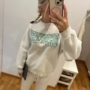 Vit sweatshirt med grön text - Vit sweatshirt med grön text 'ORIGINAL' framtill. Tröjan har rund halsringning, ribbade muddar och en relaxed passform. Perfekt för en chill och sportig look.