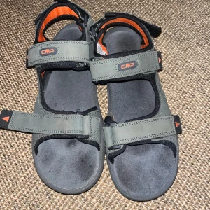 Grå sandaler från CMP med orange detaljer - Sportiga sandaler från CMP i grått med svarta och orange detaljer. Justerbara remmar med kardborre för perfekt passform. Snygg och robust design med bekväm platt sula, perfekt för sommardagar och äventyr utomhus.