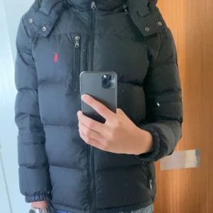 Svart dunjacka Polo Ralph Lauren L - Inga slitningar eller nåt. Svart dunjacka från Polo Ralph Lauren med huva och dragkedjor. Klassisk pufferstil med röd logga på bröstet och tre fickor med dragkedja. Perfekt för kalla dagar och riktigt snygg till streetwear. Jackan är långärmad och har en clean, stilren look.