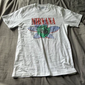 Vit Nirvana t-shirt med blommor - Vit t-shirt från Valient med stort Nirvana-tryck och färgglada blommotiv både fram och bak. T-shirten har klassisk passform, rund hals och korta ärmar. Perfekt för dig som gillar bandmerch och vill sticka ut med coola tryck.
