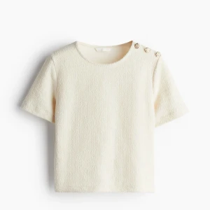 Beige bouclé topp med gulddetaljer - Snygg beige topp i bouclé-liknande material med kort ärm och rund hals. Tre dekorativa guldknappar på ena axeln ger en lyxig touch. Säljer för jag ej använder den, den är fortfarande i nyskick. 