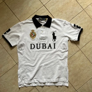 Vit pikétröja Polo Ralph Lauren Dubai - Vit pikétröja från Polo Ralph Lauren med svart krage och detaljer. Stor broderad logga, Dubai-text och emblem framtill. Snygg siffra 2 på ryggen och ärmarna. Klassisk passform och kortärmad, perfekt till en clean och sportig look.
