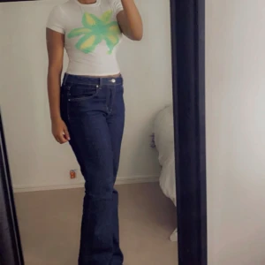 Vit t-shirt med grön/gul blomma SHEIN - Vit croppad t-shirt från SHEIN med en stor grön och gul blomma tryckt på bröstet. T-shirten har korta ärmar och en tight passform som sitter snyggt. Perfekt för dig som gillar färgglada prints och vill sticka ut lite extra.